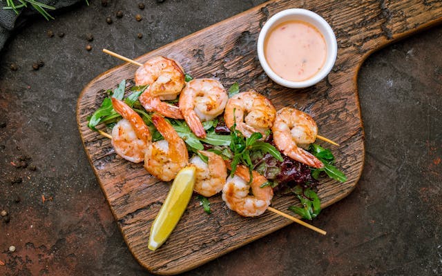 Two skewers of grilled shrimp on a bed of salad with a lemon wedge and dipping sauce